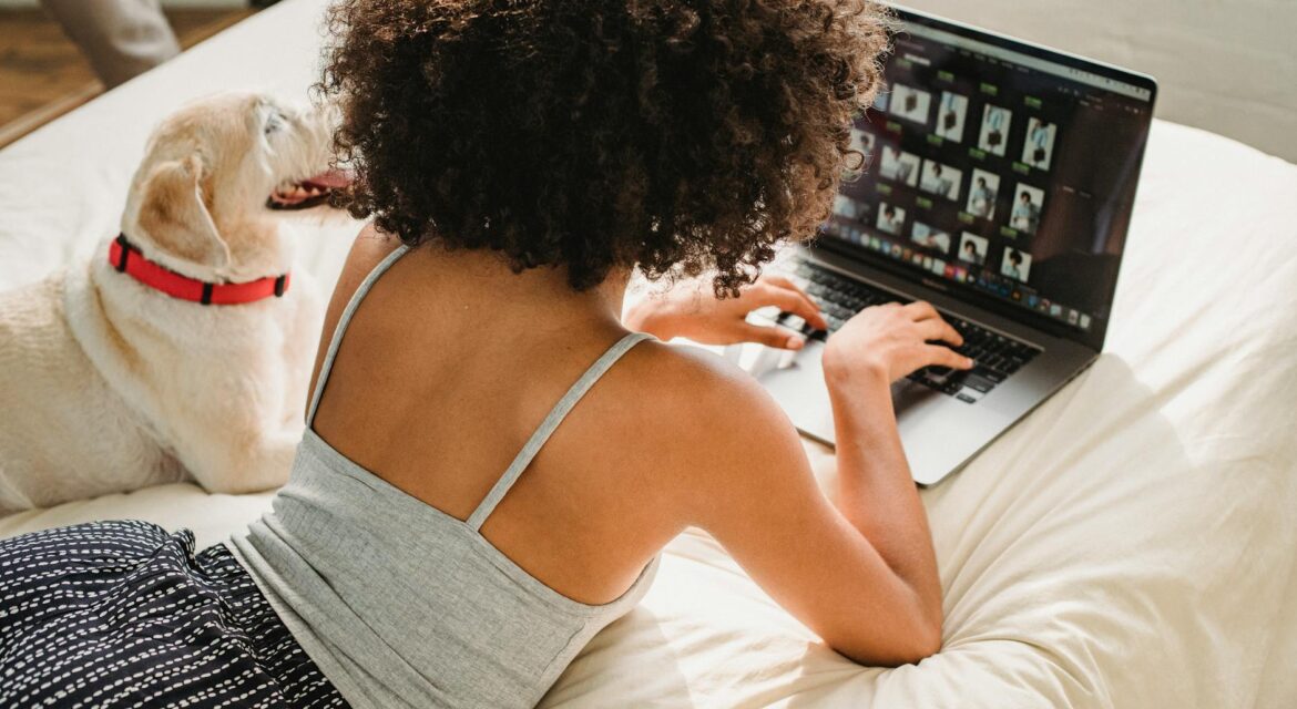 black woman browsing laptop with dog on bed